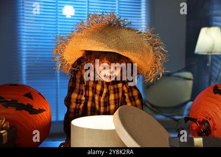 Süßer Junge, der wie Vogelscheuche gekleidet ist, mit festlichem Dekor und Geschenkboxen drinnen in der Nacht. Halloween-Feier Stockfoto