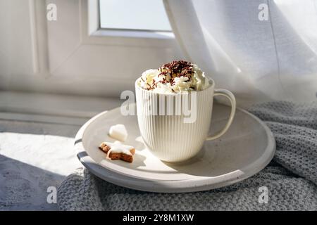 Weißer Becher mit heißer Schokolade und Schlagsahne am Fenster, warmes süßes Getränk für gemütliche Tage drinnen im Herbst und Winter, Kopierraum, ausgewähltes foc Stockfoto