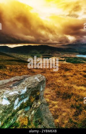 Eine große Wolke dominiert den Himmel und wirft einen Schatten über ein Tal darunter. Das Tal ist mit grasbewachsenen Hügeln und einem See gefüllt. Ein einsamer Felsen sitzt auf einem gr Stockfoto