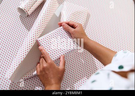 Hände, die ein Geschenk mit weißem und rotem Briefpapier umhüllen, das die Kunst des Geschenks und der Weihnachtsvorbereitung unterstreicht. Stockfoto