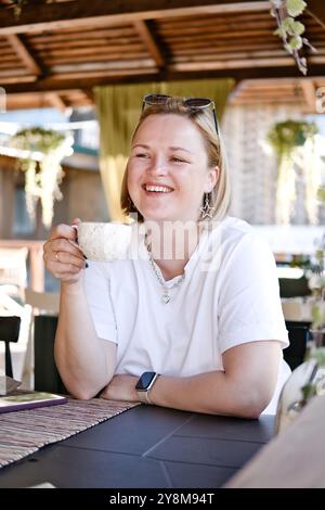 Glückliche lächelnde Frau genießt eine Tasse Kaffee oder Tee im Restaurant im Freien. Eine junge Frau, die sich beim Kaffeetrinken auf der Terrasse des Cafés entspannt. Stockfoto