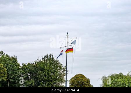 Fahnenmast mit drei Flaggen von Holland, Deutschland und blauer Dreiecksflagge, Baumkronen mit grünem Laub vor grauem Himmel im Hintergrund, bewölkter Tag in Maas Stockfoto