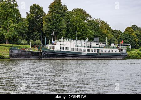 Zwei Boote ankern an einem kleinen Kai am Ufer entlang der Maas, üppige grüne Laubbäume im Hintergrund, bewölkter Tag in Maastricht, South Limburg, N Stockfoto