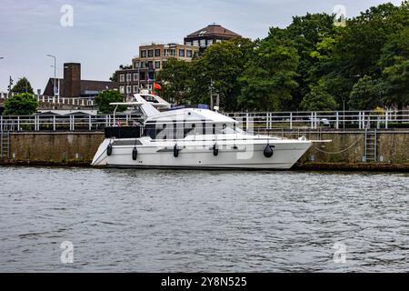 Weiße Yacht vor Anker an einem kleinen Kai am Ufer entlang des Flusses Maas, Stadtlandschaft mit Gebäuden im Hintergrund, Metallgeländer an der Promenade, bewölkter Tag Stockfoto