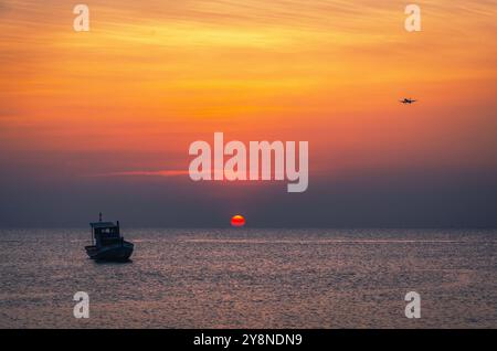 Das Flugzeug landet über einem Fischerboot, das vor dem Hintergrund eines wunderschönen Sonnenuntergangs auf den Wellen schwingt Stockfoto