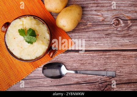 Cremiger Kartoffelpüree in einer Schüssel mit ganzen Kartoffeln und Löffel, auf einer orangen Tischdecke über einem rustikalen Holztisch, perfekt für Thanksgiving Stockfoto