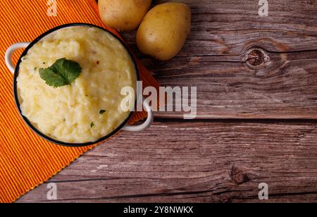 Schüssel mit Kartoffelpüree auf einem orangen Tuch, begleitet von ganzen Kartoffeln auf einem alten Holztisch. Ideal für Thanksgiving-Abendessen Stockfoto