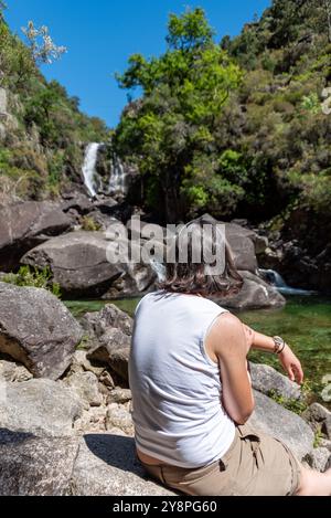 Eine junge Frau sitzt an einem Fluss und genießt den Blick auf die Rajada-Kaskade im Peneda-Geres-Nationalpark, Portugal Stockfoto