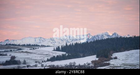 Winterlandschaft mit schneebedeckten Bergketten, Wäldern und Wiesen bei farbenprächtigem Sonnenuntergang Stockfoto