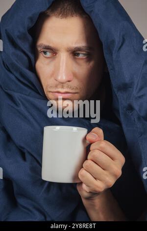 Das Bild eines jungen Mannes sitzt auf einem Bett und ist mit einer dunkelblauen Decke bedeckt. Er hält eine weiße Tasse Tee in der Hand. Der Kerl schaut in die Kamera Stockfoto