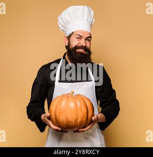 Lächelnder bärtiger Koch mit Kürbis. Männlicher Koch in Kochmütze und Schürze mit Kürbis für Halloween. Thanksgiving-Tag kochen. Herbsternte. Saisonales Vitamin Stockfoto