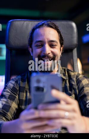 Fröhliche männliche Person, die sein Mobilgerät greift und online mit Freunden und Familie chatten. Aufgeregter junger Mann, der auf seinem Handy im Internet surft und sich lustige Inhalte in sozialen Medien ansieht. Stockfoto