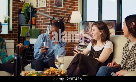 Kulturell inkludierte Gruppe von Frauen, die sich unterhalten, Wein trinken, bei Versammlungen, tratschen über ihre Freunde. Asiatische, afroamerikanische und kaukasische Mädchen, die das soziale Leben im Wohnzimmer genießen Stockfoto