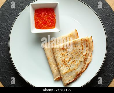 Dünne Pfannkuchen mit rotem Kaviar auf weißem Teller, die auf schwarzer texturierter Tischmatte liegen Stockfoto