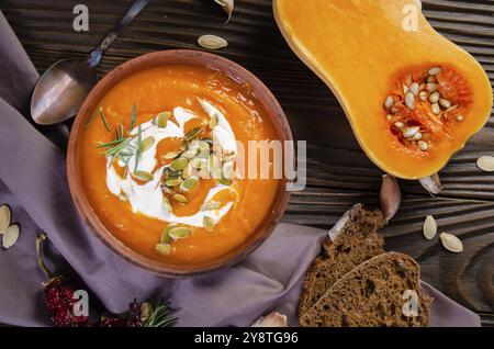 Blick auf die hausgemachte rustikale Kürbissuppe mit Samen in Tonform auf Holztisch mit Brot-Gewürzen und Grüns Stockfoto