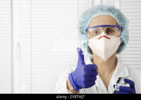 Porträt der glücklich lächelnden kaukasischen Ärztin in Maske Brille Handschuhe und Hut zeigt Daumen auf weißem Fensterhintergrund Stockfoto