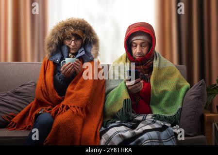 Mittelalte Familie in Parkas, die Kaffee auf dem Sofa trinkt, während sie das Smartphone in ihrem kalten Wohnzimmer benutzt Stockfoto