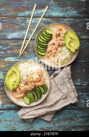 Zwei weiße Keramikschalen mit Reis, Garnelen, Avocado, Gemüse, Sesamsamen und Essstäbchen auf farbenfrohem rustikalem Holzhintergrund von oben. Gesundheit Stockfoto