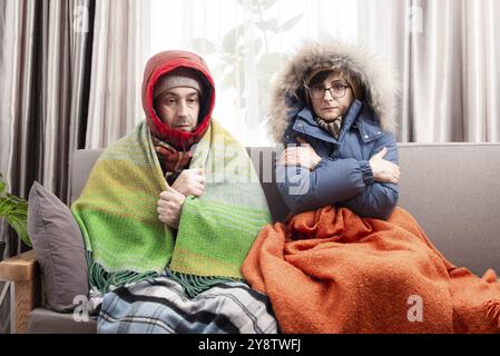 Trauriges Paar mittleren Alters in warmen Kleidern, das auf dem Sofa in ihrem Wohnzimmer kalt wird Stockfoto