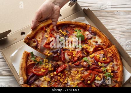 Menschliche kaukasische Hand nimmt ein Stück große Pizza aus der Schachtel auf weißem Holztisch Stockfoto