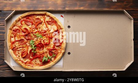 Peperoni Pizza mit Paprika und Rucola in offener Kartonbox auf dunklem Holztisch flach liegend mit Kopierraum Stockfoto