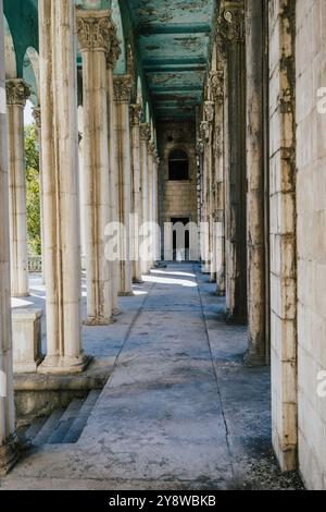 Verlassener Korridor mit hohen, verwitterten Steinsäulen und abblätternder blauer Decke, der zu einem dunklen Eingang führt. Stockfoto