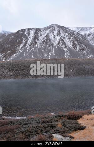 Die Verdunstung steigt über der Oberfläche eines breiten Sees mit hohen felsigen Ufern im ersten Herbstschnee an einem bewölkten Tag. Kok-Kol-See, Altai, Sibirien, Russ Stockfoto