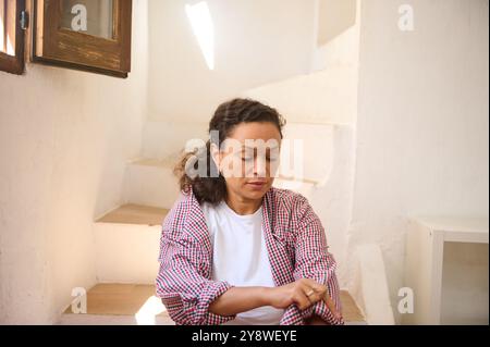 Eine Frau in einem karierten Hemd sitzt nachdenklich auf einer sonnendurchfluteten Indoor-Treppe und strahlt ruhige Reflexion aus. Stockfoto