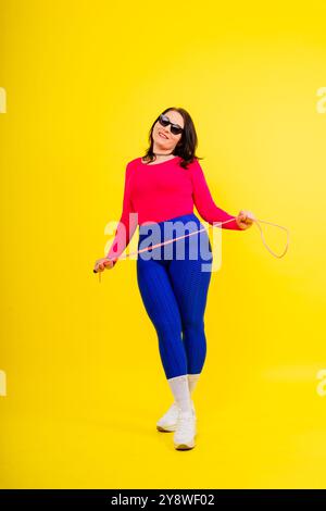 Hübsch lächelnde, pralle Frau in einem sportlichen Oberteil und Leggings, die ein Springseil in den Händen hält Stockfoto