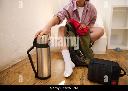 Person, die sich auf eine Wanderung vorbereitet, packt Essentials wie eine Thermosflasche und ein erste-Hilfe-Set. Konzentrieren Sie sich auf Vorbereitung und Organisation in einem gemütlichen Wohnhaus Stockfoto