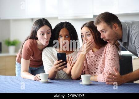 Vier schockierte interrassische Freunde, die zu Hause in der Küche telefonieren Stockfoto