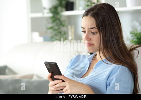 Enttäuschte Frau überprüft das Smartphone, das zu Hause auf einer Couch sitzt Stockfoto