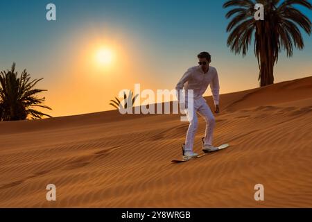 Glückliche männliche Touristen Sandboarding auf Sanddünen in der Wüste während Sonnenuntergang Stockfoto