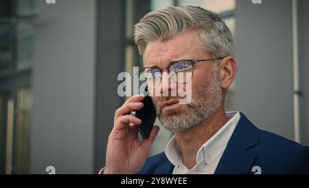 Ernsthafter erwachsener Unternehmer mittlerer Alter Geschäftsmann in der Stadt in der Nähe von Geschäftsgebäuden verwirrte Antwort Anruf aufgebracht besorgt Brille abzunehmen Senior Stockfoto