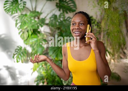 Zuversichtlich zufriedene Afroamerikanerin, die am Telefon spricht, umgeben von grünem Minigarten Stockfoto