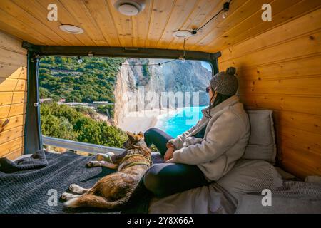 Frau und Hund auf dem Bett des Wohnwagens und schauen auf den Strand mit türkisfarbenem Wasser Stockfoto