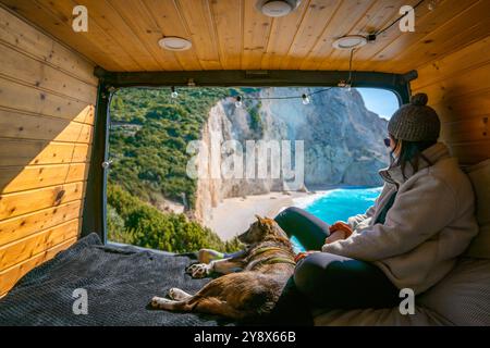 Frau und Hund auf dem Bett des Wohnwagens und schauen auf den Strand mit türkisfarbenem Wasser Stockfoto