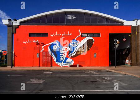 Ein leuchtend rotes Gebäude mit einem großen blauen Sneaker-Wandgemälde vor einem klaren blauen Himmel in der O'Village Mall in casablanca Stockfoto