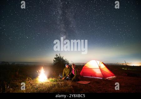 Ein paar Reisende sitzen in der Nähe eines beleuchteten Zeltes unter einem wunderschönen Himmel mit Sternen. Nächtlicher Sternenhimmel über einem grasbewachsenen Hügel mit Touristenzelt, Lagerfeuer und Wanderern. Konzept von Wandern und Nachtcamping. Stockfoto