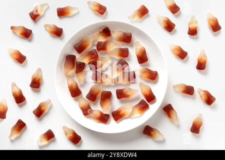Gummiartige Gelees in Form von Soda-Flaschen und voller weißer Süßigkeiten-Platte auf weißem Hintergrund. Blick von oben. Stockfoto