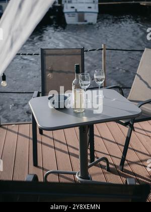 Eine Flasche Weißwein und zwei Gläser auf einem modernen Hausboot mit ruhigem Wasser und Sonnenschein mit Lichtern und Booten im Hintergrund Stockfoto