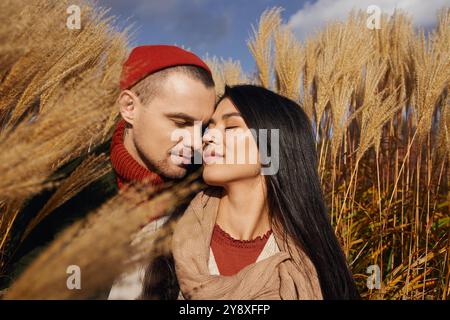 Ein Paar, das warm gekleidet ist, umarmt sich und lächelt sanft vor einem Hintergrund aus Herbstlaub und Gräsern. Stockfoto