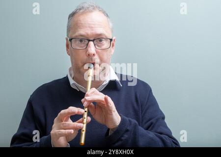 Mann mit grauen Haaren, lässig gekleidet, mit dunkler Brille, spielt eine goldfarbene Blechpfeife, Pennypfeife, traditionelle irische Volksmusik. Stockfoto