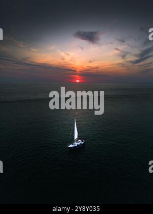 Ein ruhiges Segelboot auf dem offenen Meer bei Sonnenuntergang, mit dramatischem Himmel und einem leuchtenden orangefarbenen Sonnenuntergang über dem Horizont. Stockfoto