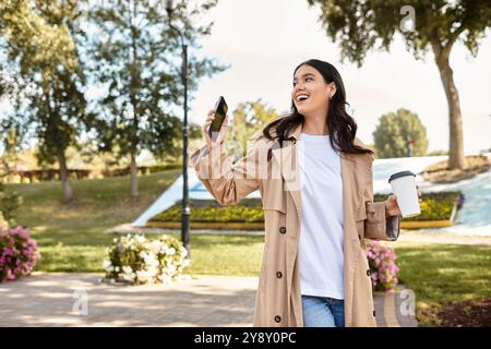 Eine fröhliche Frau, die im Herbst gekleidet ist, schlürft durch einen lebhaften Park, schlürft Kaffee und lächelt. Stockfoto