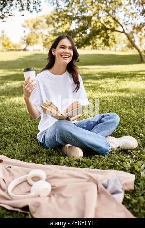 Eine lächelnde Frau sitzt auf dem Gras und trinkt Kaffee, während sie ihr Buch in einem ruhigen Herbstpark liest. Stockfoto