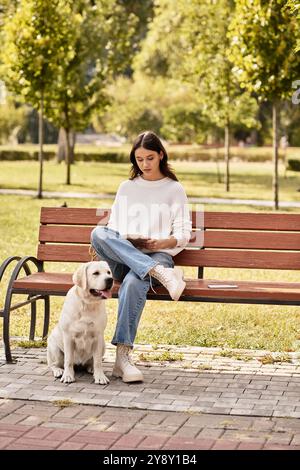 Eine schöne Frau in kuscheliger Herbstkleidung sitzt auf einer Bank und liest mit ihrem Hund an ihrer Seite. Stockfoto