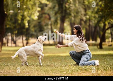 Eine schöne Frau in kuscheliger Kleidung kniet im Park und wirft ihrem energiegeladenen Hund einen Ball zu. Stockfoto