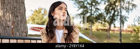 Eine junge, schöne Frau sitzt besinnlich auf einer Parkbank, umgeben von Herbstlaub. Stockfoto