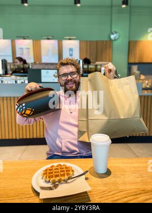 Ein überraschter Mann sitzt mit einer Waffel an einem Café-Tisch, hält eine große Einkaufstasche und eine Geschenkbox zum Mitnehmen. Moment der unerwarteten Freude und des Genusses in einem Stockfoto